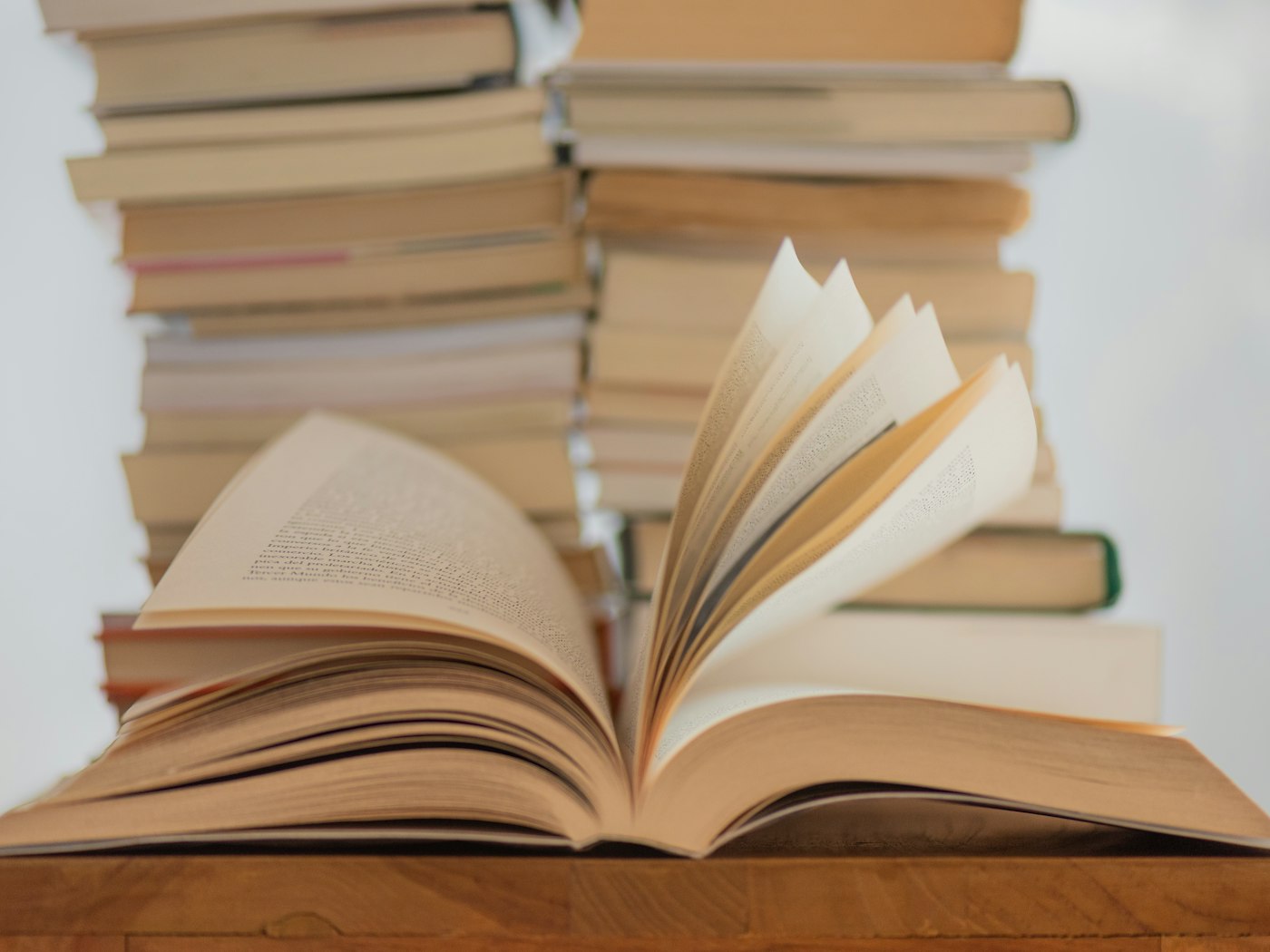 An open book resting on a wooden table in front of tall stacks of books