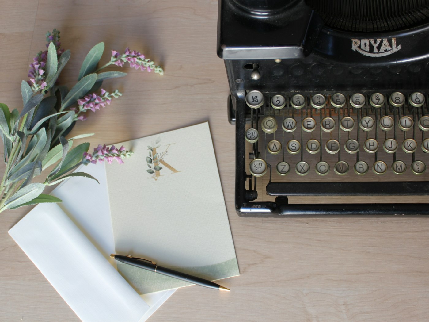 A vintage black Royal typewriter beside cream stationery with a botanical letter K, a black and gold pen, and sprigs of lavender on pale wood