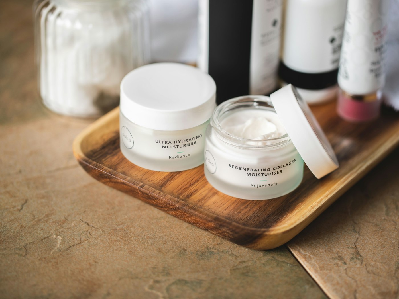 Two frosted glass jars of moisturizer on a wooden tray against a warm stone background